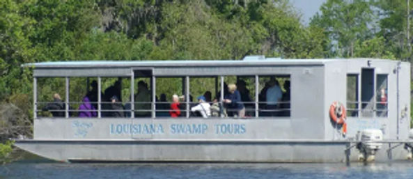 Open-sided tour boat with seated passengers cruising a Louisiana bayou, metal cabin and orange life ring visible, lush green cypress shoreline in the background.