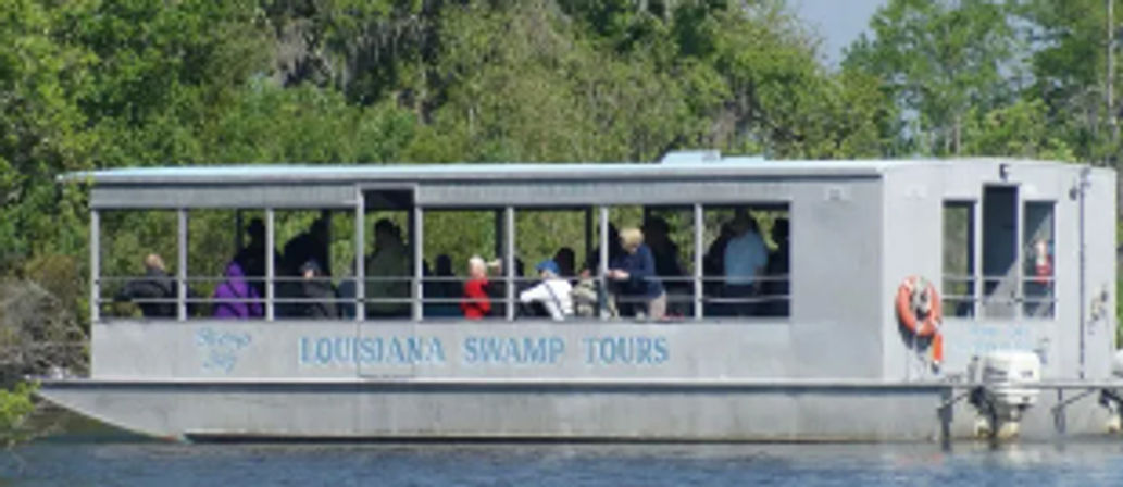 Open-sided tour boat with seated passengers cruising a Louisiana bayou, metal cabin and orange life ring visible, lush green cypress shoreline in the background.