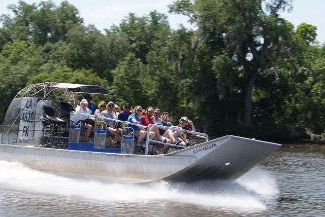 Passengers zooming on an airboat through a Louisiana bayou, water spray trailing past moss-draped cypress trees and lush green shoreline.