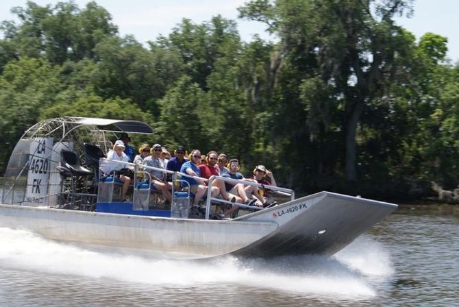 Passengers zooming on an airboat through a Louisiana bayou, water spray trailing past moss-draped cypress trees and lush green shoreline.