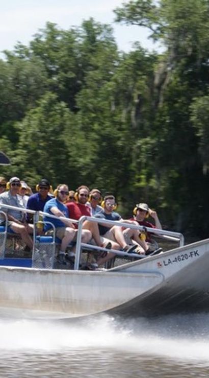 Passengers zooming on an airboat through a Louisiana bayou, water spray trailing past moss-draped cypress trees and lush green shoreline.