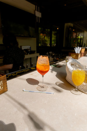 Sunlit outdoor bar counter with a bright orange iced cocktail in a stemmed glass casting a shadow, a second glass of yellow iced juice, a wrapped straw and a woven white bag on a pale stone countertop.