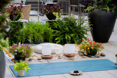 Outdoor urban patio meditation setup with blue yoga mat, round floor cushion, two crystal singing bowls on wooden stands, colorful potted flowers, ferns and decorative stones creating a zen garden vibe.