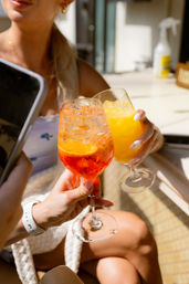 Sunlit brunch scene on an outdoor patio: two hands clinking stemmed glasses—one a bright orange aperitif with ice and citrus, the other a yellow-orange juice cocktail.
