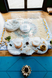 Sunlit indoor meditation setup by glass doors on a hardwood floor: round cushions on a mandala blanket, crystal singing bowls, healing crystals, tassels, small plants and a blue yoga mat.
