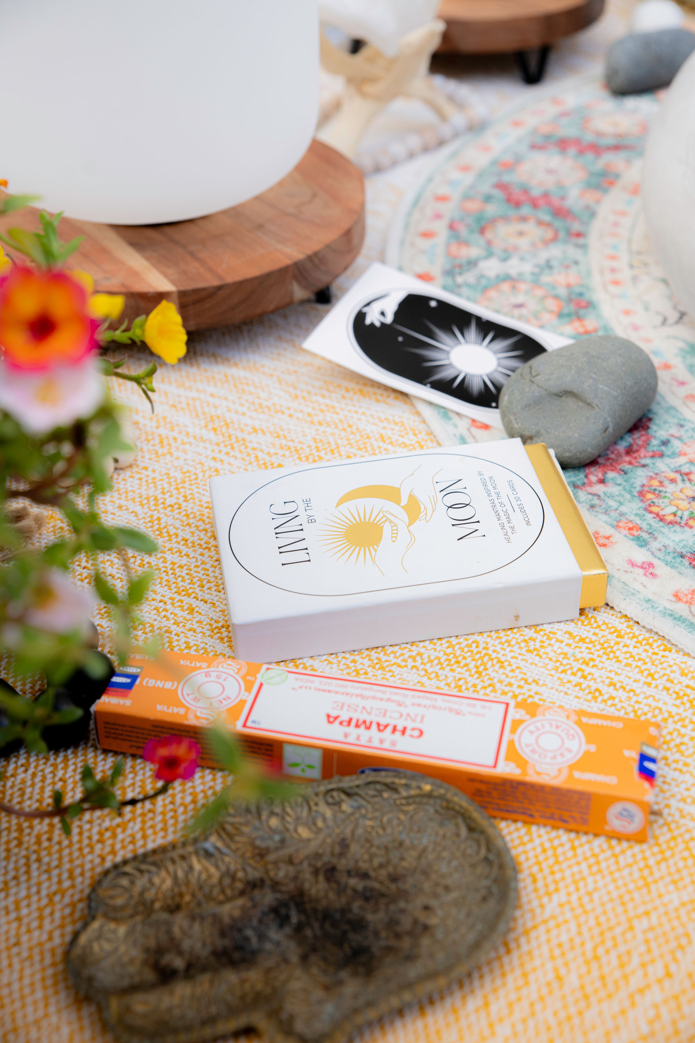 Boho home altar flatlay with a moon-themed oracle card deck, incense sticks, smooth ritual stones, a crystal singing bowl on a wooden stand, colorful patterned rug and fresh flowers.