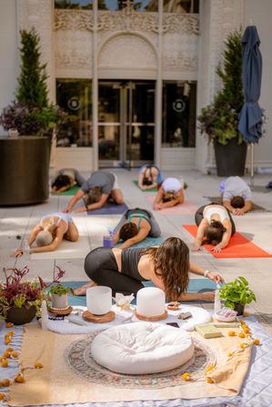 Outdoor yoga class on a sunlit urban plaza: group in child's pose on colorful mats in front of an ornate building, with meditation cushions, crystal singing bowls, potted plants and wellness props in the foreground.