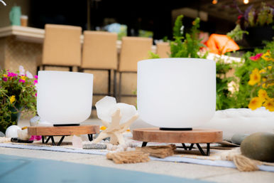 Two frosted crystal singing bowls on wooden stands surrounded by crystals and colorful flowers on a garden patio, ready for a soothing outdoor sound bath.