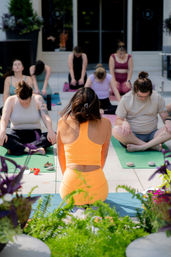 Sunlit rooftop yoga session with an instructor in a bright orange set leading a mixed group seated on mats for meditation amid potted plants on an urban patio