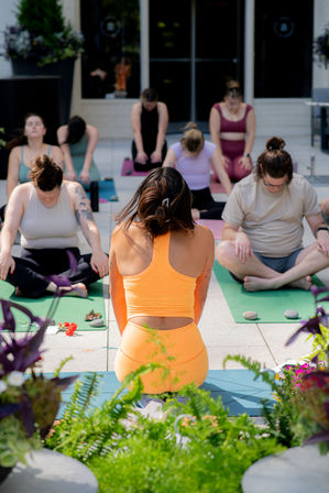 Sunlit rooftop yoga session with an instructor in a bright orange set leading a mixed group seated on mats for meditation amid potted plants on an urban patio