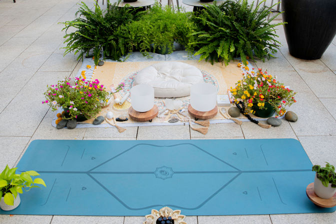 Outdoor patio meditation setup with a blue yoga mat, round white floor cushion, two crystal singing bowls on wooden stands, colorful potted flowers and ferns, and decorative stones and beads.