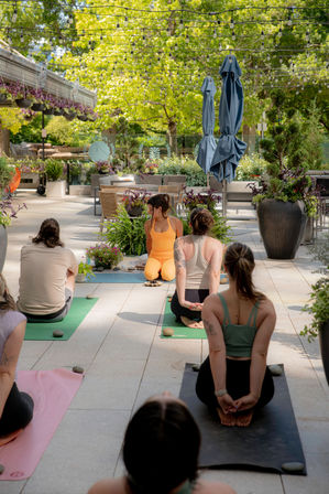 Outdoor urban garden yoga class on a sunny patio — instructor in orange leads students on mats under hanging string lights, surrounded by large planters, umbrellas, and trees.