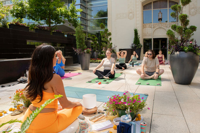 Sunny downtown outdoor terrace group yoga class with an instructor in orange leading students on colorful mats among large planters and a city building backdrop.