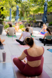 Outdoor rooftop yoga class on a sunny leafy patio — participants on mats, woman in maroon activewear with a bun in the foreground