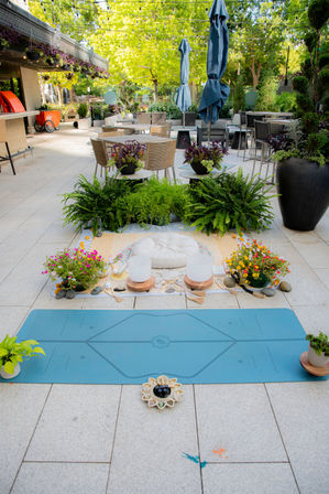 Sunlit urban garden patio with a blue yoga mat and meditation setup — round cushion, crystal singing bowls and colorful potted plants — surrounded by dining tables, umbrellas and string lights.