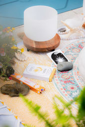 Poolside meditation setup with a white crystal singing bowl on a wooden stand, tarot card, healing stones, incense boxes and a colorful boho round rug