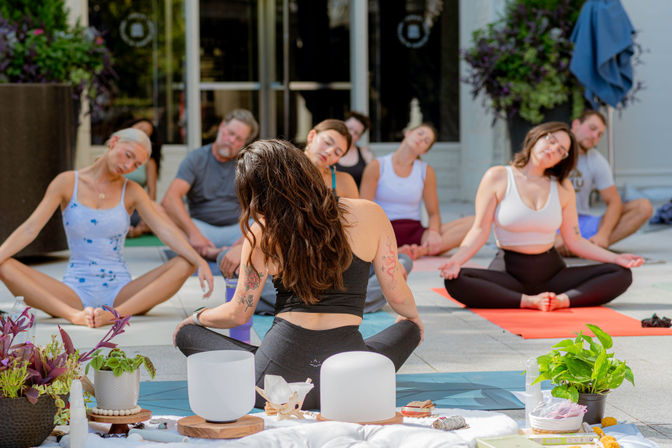 Sunlit outdoor yoga and meditation class in a courtyard — students on colorful mats stretching while an instructor leads beside singing bowls and potted plants.