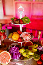 Three-tier white serving stand centerpiece piled with strawberries, kiwi slices, bananas, oranges and dates, accented by pink and purple dahlias on a festive party table with warm pink lighting.