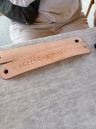 Two tan leather snap bracelets stamped with decorative icons and snaps resting on a beige linen tablecloth, a person in light clothing blurred in the background.