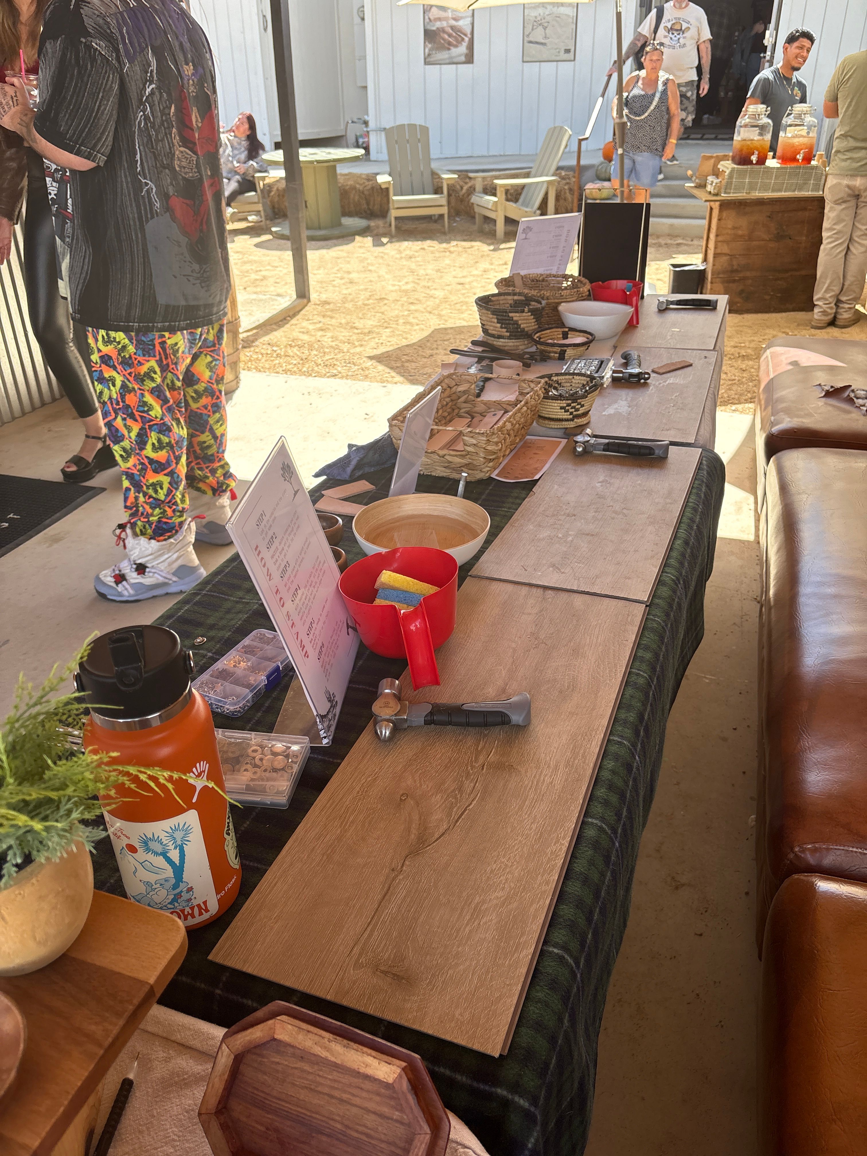 Sunny outdoor market booth displaying wood plank samples, small hammers, woven baskets and bowls arranged on a long table, with shoppers and Adirondack chairs on a sandy patio in the background.