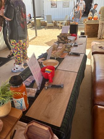 Sunny outdoor market booth displaying wood plank samples, small hammers, woven baskets and bowls arranged on a long table, with shoppers and Adirondack chairs on a sandy patio in the background.