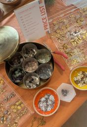Overhead shot of a DIY jewelry stall: round tin with compartments of pastel gemstone teardrop charms, clear organizers of gold metal charms and tiny connectors, bowls of silver letter and star charms, red-handled pliers and a small price list on an orange tablecloth.