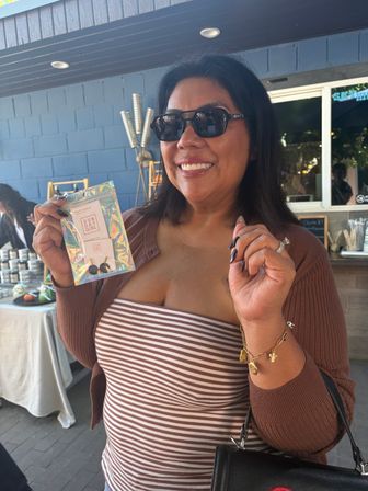 Smiling shopper in sunglasses at an outdoor market holding holographic-packaged handmade earrings with a small label, wearing a striped tube top, brown cardigan and gold charm bracelet.