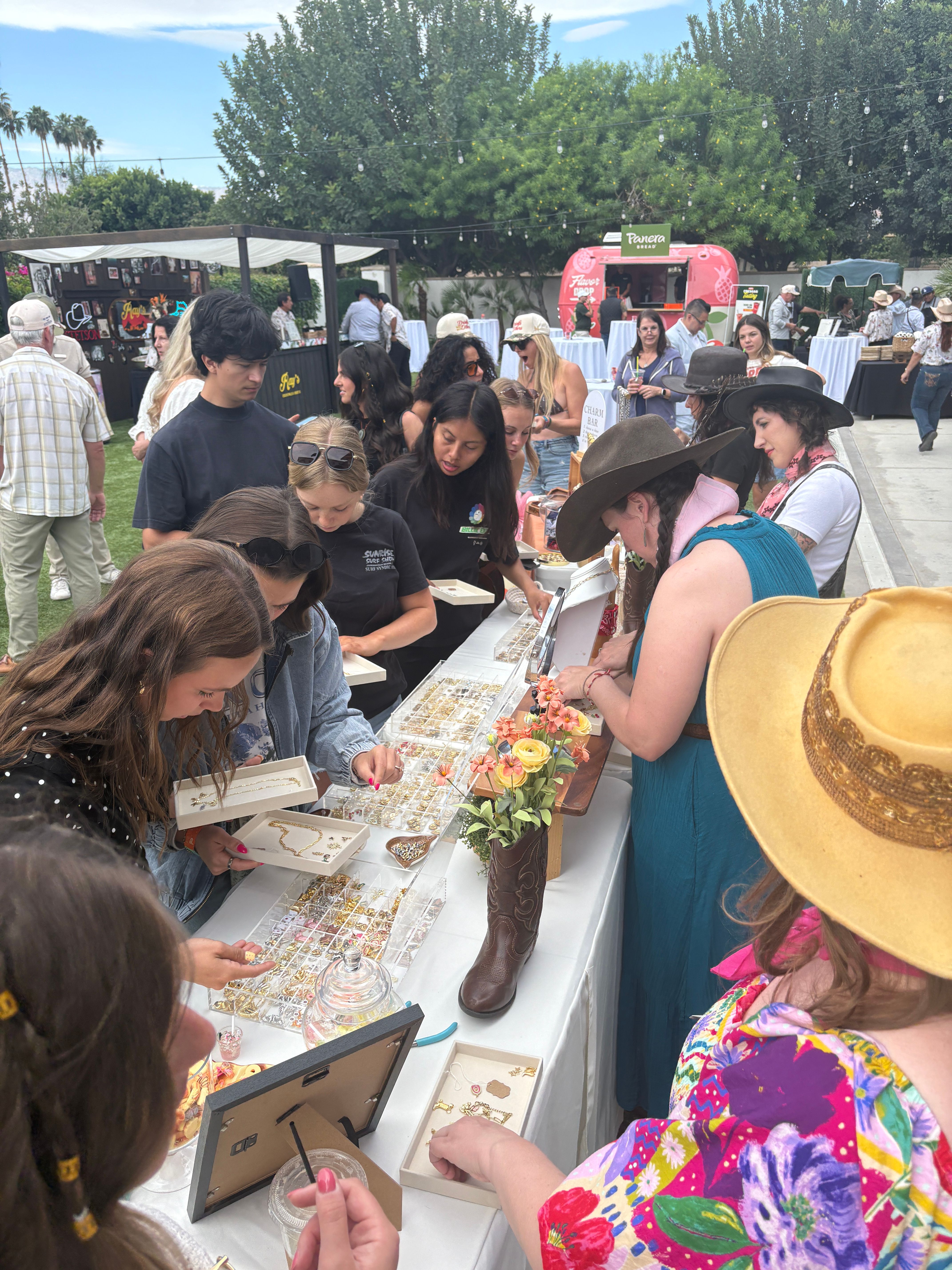 Outdoor pop-up market with shoppers crowding a jewelry vendor table, trays of gold-tone necklaces and rings, a single cowboy boot display, people in sun and western hats, string lights overhead and a pink food truck near leafy trees.