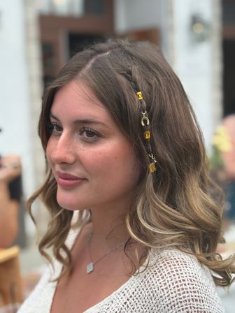 Close-up portrait of a woman with braided brown hair adorned with gold beads and a horseshoe charm, wearing a white knit top and pendant necklace, beachy summer vibe