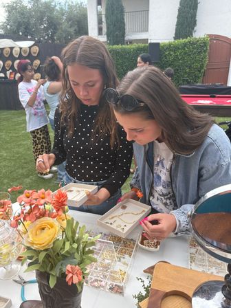 Two women leaning over trays of gold necklaces and charms at an outdoor pop-up jewelry table, floral centerpiece in the foreground, hats on a fence and guests on a backyard lawn.