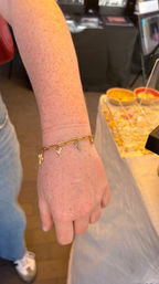 Close-up of a freckled forearm wearing a gold chain charm bracelet with cactus, martini and cross charms, shown over a jewelry display at a market booth.