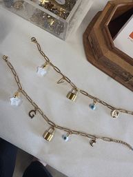 Gold chain charm bracelets with padlock, evil eye, star and moon charms displayed on cream linen beside a wooden tray and clear container of extra charms — boutique jewelry display