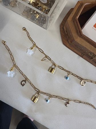 Gold chain charm bracelets with padlock, evil eye, star and moon charms displayed on cream linen beside a wooden tray and clear container of extra charms — boutique jewelry display