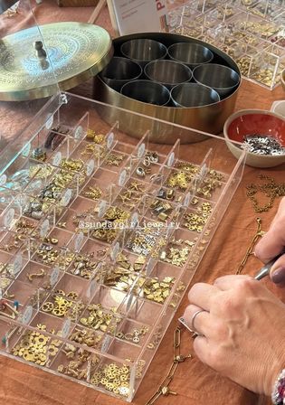 Close-up of a jewelry-making workspace: a clear organizer filled with tiny gold charms and findings, metal tins and bowls, and hands using pliers to assemble a delicate gold chain on a peach fabric-covered table.