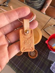 Hand holding a tan handmade leather keychain embossed with a stylized thunderbird and sunburst, attached to a metal key ring, with wooden bowls and craft supplies on a table below.