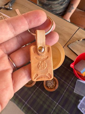 Hand holding a tan handmade leather keychain embossed with a stylized thunderbird and sunburst, attached to a metal key ring, with wooden bowls and craft supplies on a table below.