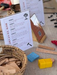 Leather stamping workshop table at an artisan market with a step-by-step instruction card, wicker basket of leather scraps, blue sponge, yellow stamping tool and embossed leather strips ready for crafting.