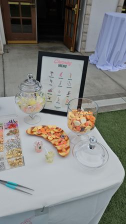 Outdoor event candy table on a patio — glass apothecary jars and large goblet of pastel sweets, wooden tray of mini treats, framed “Charming Menu” sign and tongs on a white tablecloth.