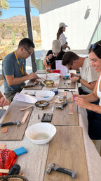 Group DIY leather-stamping workshop at an outdoor table with hammers, tools and bowls, smiling participants and a Joshua Tree desert backdrop