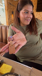 Smiling woman in an olive shirt seated in a cozy wood‑paneled café booth, holding and showing a stamped leather key fob with stars, a smiley and letters.