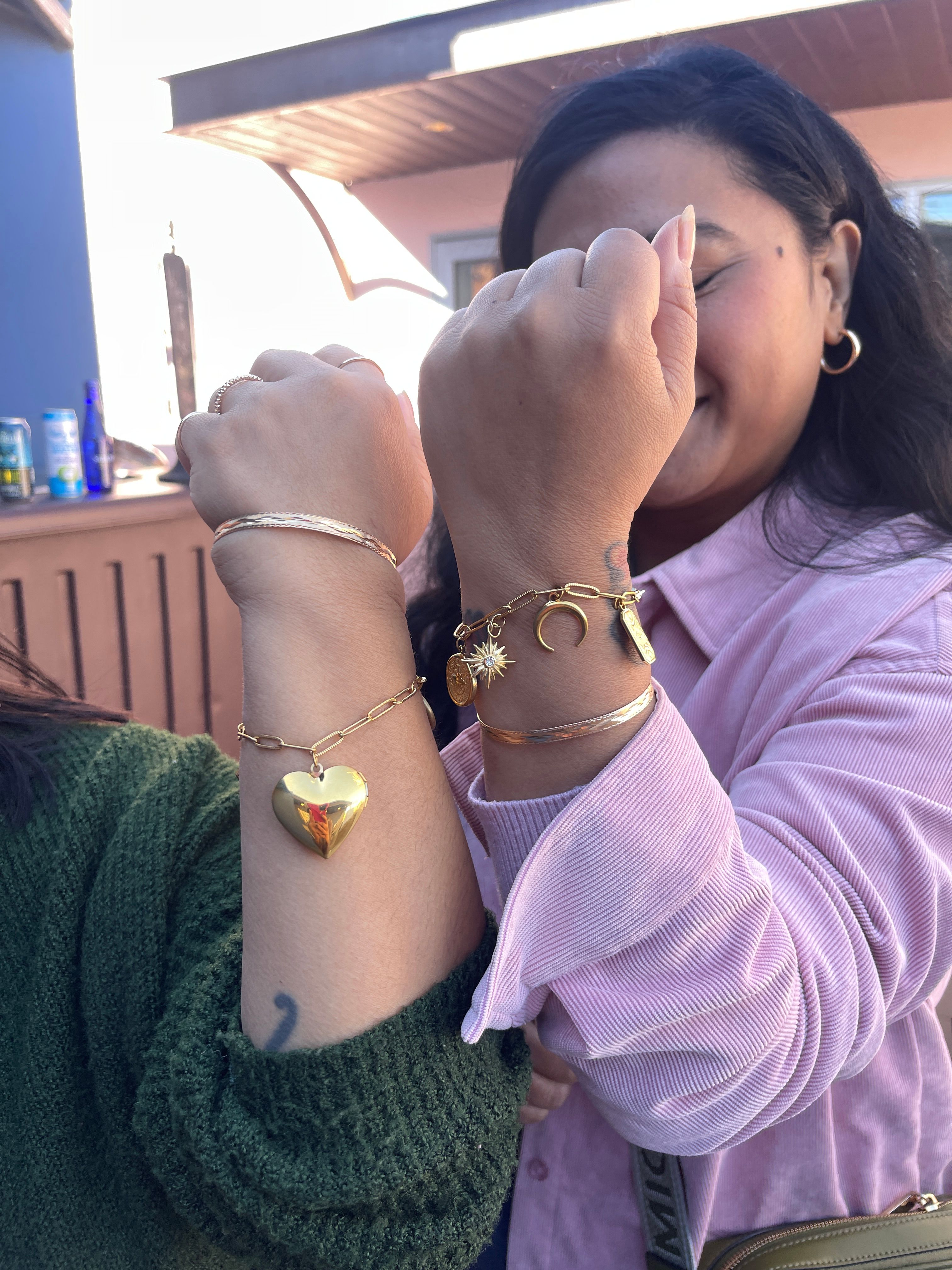 Two friends on an outdoor patio hold up wrists to show gold charm bracelets and bangles — heart locket pendant, crescent moon and sun charms — one in a pink corduroy shirt and one in a green knit sweater.