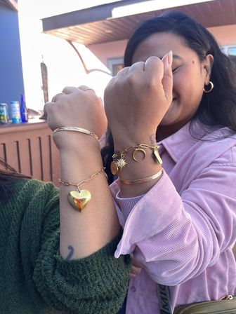 Two friends on an outdoor patio hold up wrists to show gold charm bracelets and bangles — heart locket pendant, crescent moon and sun charms — one in a pink corduroy shirt and one in a green knit sweater.
