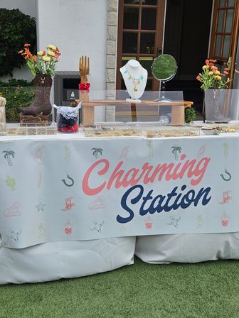 Outdoor craft-fair vendor table showcasing jewelry and accessories: mannequin bust with colorful necklaces, wooden display hand with bracelets, round mirror, clear jewelry trays, floral arrangements in cowboy-boot vases and a bright banner across the front.