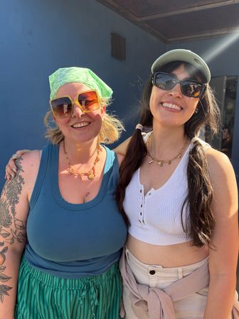 Sunlit outdoor portrait of two smiling friends in sunglasses and summer outfits posing against a blue wall — one with a green bandana and yellow shades, the other in a cap and black sunglasses.