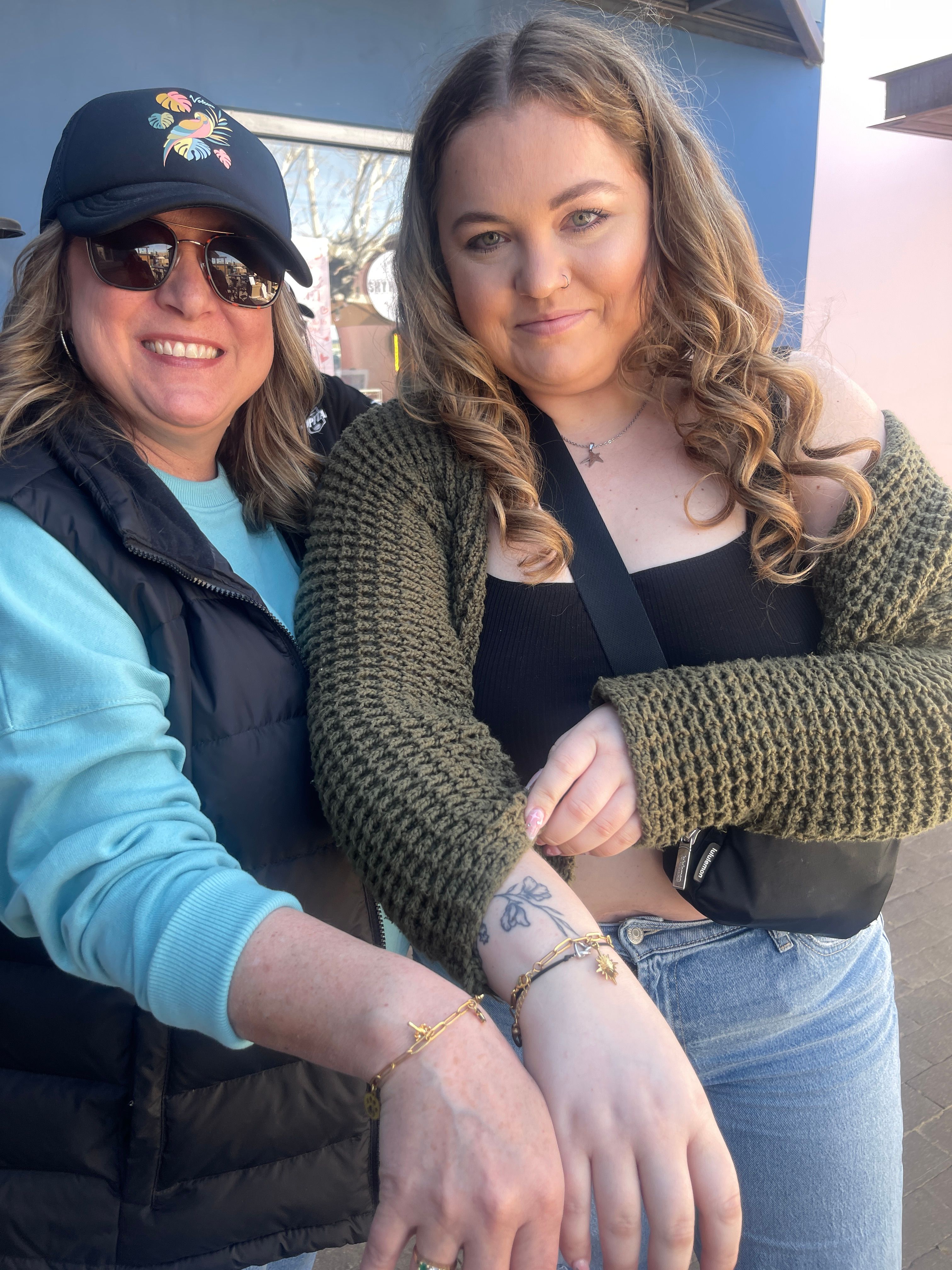 Two smiling friends outdoors on a sunny main street showing matching gold charm bracelets and a small floral wrist tattoo, wearing casual knitwear and sunglasses.