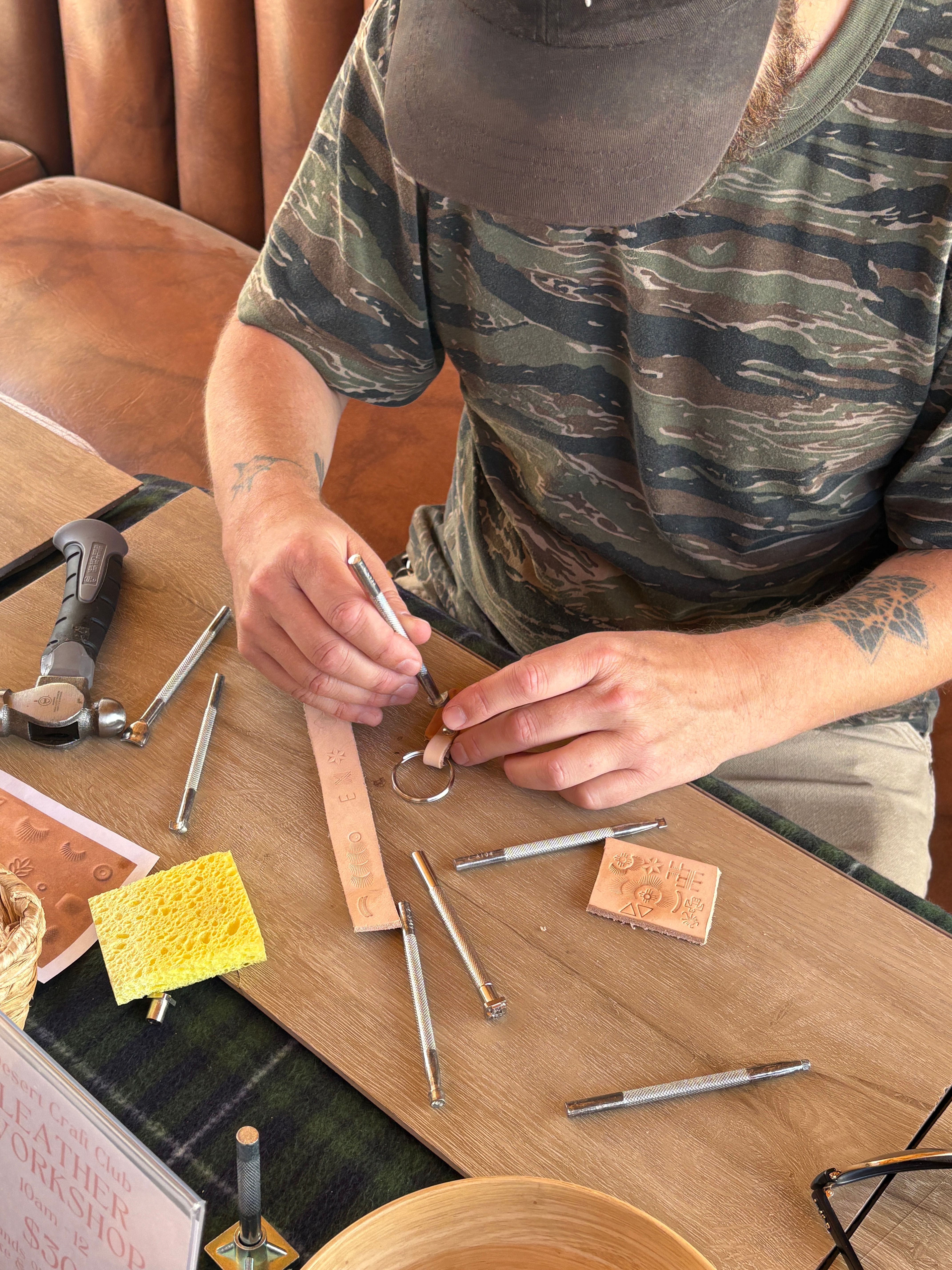 Close-up of hands doing leathercraft: stamping a leather keychain on a wooden workbench with metal stamps, hammer, sponge and scraps scattered, person wearing camo shirt at a workshop