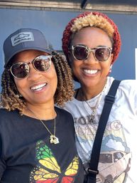 Two smiling women outdoors wearing sunglasses and layered necklaces, one in a butterfly graphic tee and cap, the other in a car-print shirt and red knit beanie, posing close together.