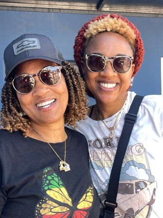 Two smiling women outdoors wearing sunglasses and layered necklaces, one in a butterfly graphic tee and cap, the other in a car-print shirt and red knit beanie, posing close together.
