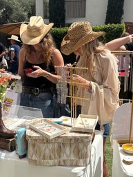 Two women in straw cowboy hats browsing gold and silver necklaces at a sunny outdoor pop-up market jewelry stall