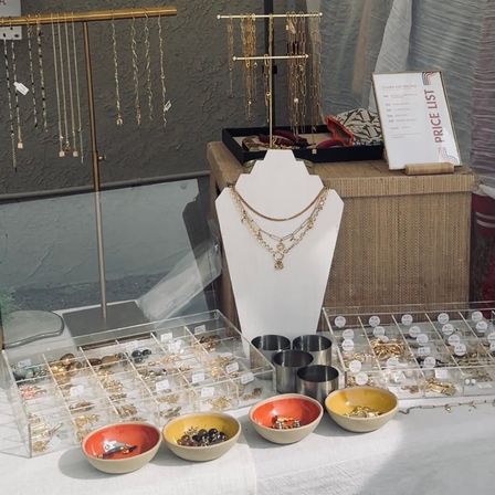 Artisan market jewelry display at an outdoor booth with gold necklaces on a white bust, hanging chain stands, clear trays of rings and earrings, and small colorful bowls of charms with a tabletop price list